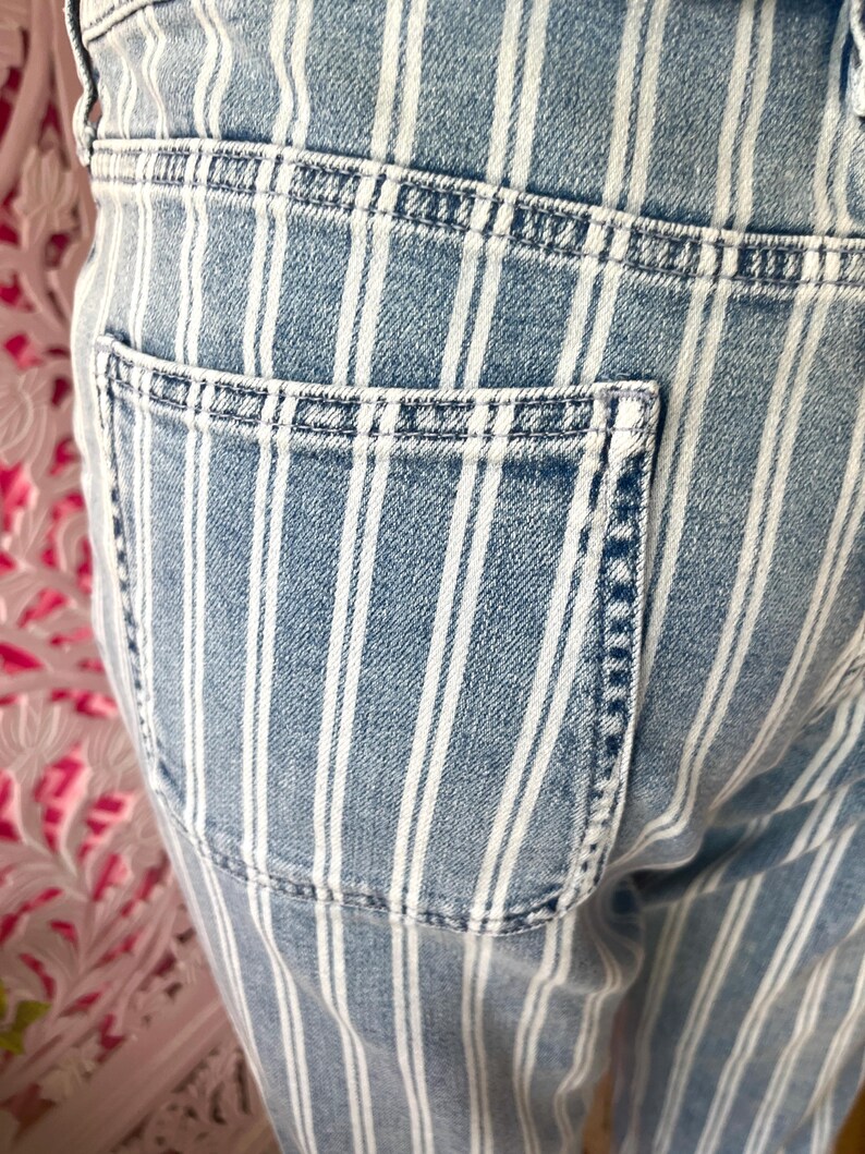 Pode incluir: Close-up de jeans de ganga azul claro com riscas brancas verticais. Os jeans apresentam um bolso traseiro e detalhes de costura vis&iacute;veis. O tecido tem um aspeto desbotado e gasto.