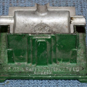 May include: A green metal ashtray with a silver top. The ashtray has a rectangular base with a raised design and the text "CENTRAL PATTERN & FOUNDRY CO. CHICAGO" on the front.