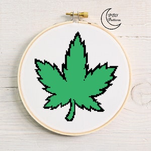 May include: A green marijuana leaf cross stitch pattern in a white embroidery hoop. The pattern is in a pixelated style.