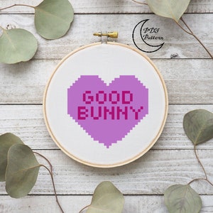 May include: A purple heart-shaped cross stitch pattern with the text "GOOD BUNNY" in pink. The pattern is displayed in a wooden embroidery hoop on a white wooden background.