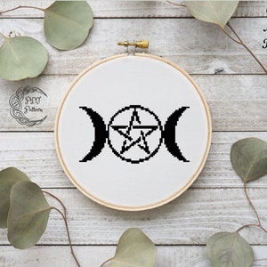 May include: A black and white cross stitch pattern of a triple moon symbol with a pentagram inside the centre moon. The pattern is on a white fabric and is in a wooden embroidery hoop.