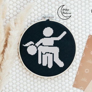 May include: A black cross-stitch embroidery hoop featuring a white pixelated design of two figures. The text "Celtic Moon Black" is displayed in white. The hoop is set against a neutral textured background.