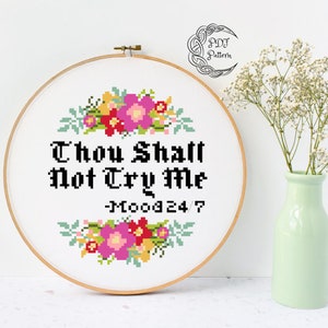May include: A white embroidery hoop with a cross-stitch design featuring a floral border and the text "Thou Shalt Not Try Me -MOOD 24/7".