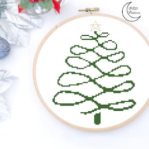 Peut inclure: Un motif de nœud celtique vert en forme de sapin de Noël avec une étoile dorée au sommet. Le motif est sur un fond blanc dans un cerceau de broderie en bois.