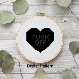 May include: Black and white cross-stitch pattern of a heart with the text "F*CK OFF" inside. The pattern is displayed in a white embroidery hoop on a wooden surface.