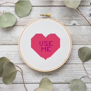 May include: A pink heart-shaped cross-stitch pattern with the text "USE ME" in purple. The pattern is displayed on a white embroidery hoop against a wooden background.