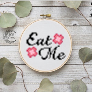 May include: A white embroidery hoop with black text that reads "Eat Me" and three pink flower designs. The hoop is on a wooden background.