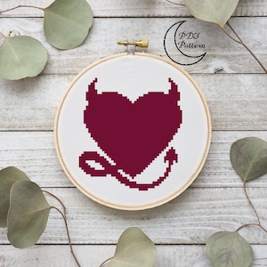 May include: A burgundy heart with devil horns and a tail, cross-stitched on white fabric in a round embroidery hoop. The hoop is on a white wooden surface.