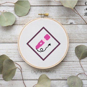 May include: A pink and black cross stitch pattern of a Celtic moon symbol in a square frame. The pattern is displayed on a white embroidery hoop.