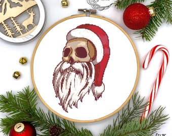 May include: Cross stitch pattern of a skull wearing a Santa hat and a white beard. The pattern is displayed in a wooden embroidery hoop on a white background.