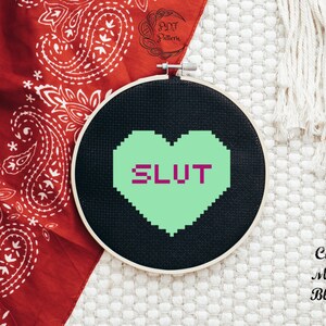 May include: A black and green cross-stitch embroidery hoop with a green heart that says "SLUT" in purple letters.