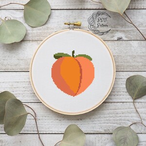 May include: A cross-stitch embroidery hoop featuring a peach design. The peach is a pixelated design with a green stem and leaf. The hoop is on a white wooden background.