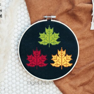 May include: A cross-stitch pattern featuring three stylized maple leaves in shades of green, red, and yellow on a black background. The pattern is displayed in a wooden embroidery hoop. The text "Celtic Moon Black" and "Digital Pattern" are visible below the hoop.