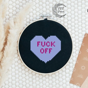 May include: A black embroidery hoop with a purple heart that says "F*CK OFF" in pink letters. The hoop is on a white textured surface.