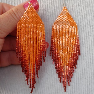 May include: A pair of orange and red beaded earrings with a fringe design. The earrings are made with small, square beads that are arranged in a gradient pattern, starting with a light orange at the top and fading to a deep red at the bottom. The earrings are suspended from silver hooks.