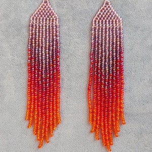 Puede incluir: Un par de pendientes largos de cuentas con un degradado de cuentas moradas, rojas y naranjas. Los pendientes cuelgan de ganchos plateados.