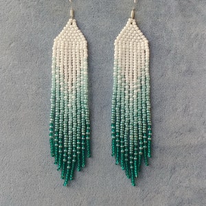 May include: A pair of long, beaded earrings with a white to green ombre design. The earrings are made with small, round beads and have a silver hook closure.