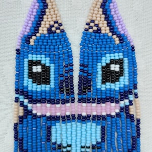 May include: Handmade beaded earrings in a whimsical design. The earrings feature a blue, pink, and black face-like pattern with dangling fringe. The earrings are made with small beads and have silver-colored ear wires. The artist's name is visible.