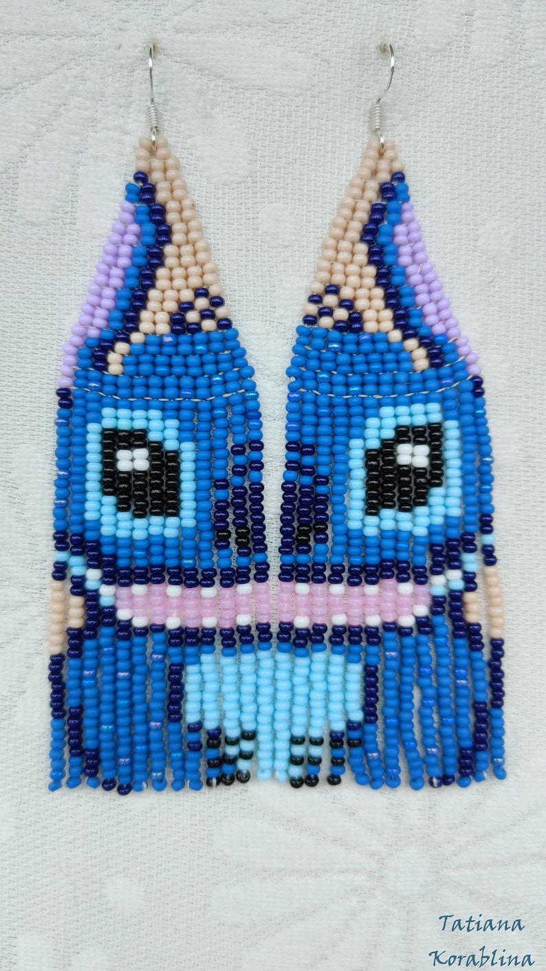 May include: Handmade beaded earrings in a whimsical design. The earrings feature a blue, pink, and black face-like pattern with dangling fringe. The earrings are made with small beads and have silver-colored ear wires. The artist's name is visible.