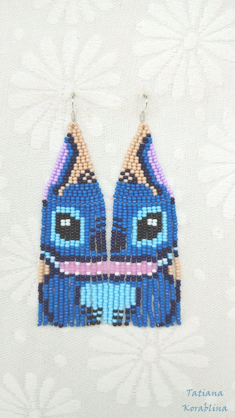 May include: Handmade beaded earrings featuring a blue cartoon character design. The earrings have a blue, black, pink, and light blue color scheme. The earrings are approximately 3 inches long.