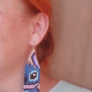 May include: Blue beaded earrings with a cartoon character design. The earrings are rectangular with a fringe at the bottom. The design includes black, white, pink, and light blue beads. The earrings are hanging from a silver hook.