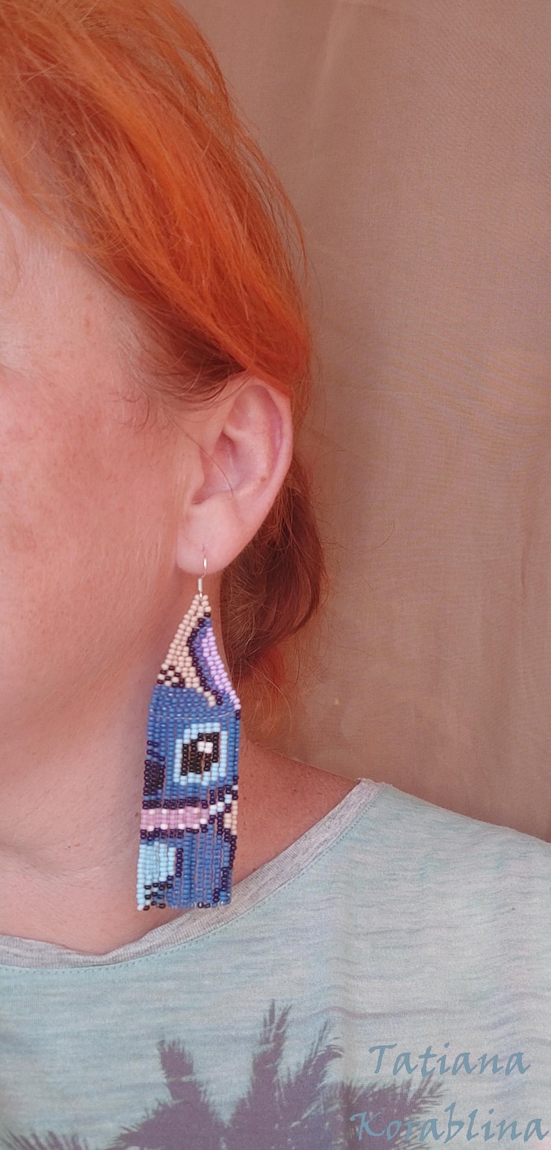 May include: Blue beaded earrings with a cartoon character design. The earrings are rectangular with a fringe at the bottom. The design includes black, white, pink, and light blue beads. The earrings are hanging from a silver hook.