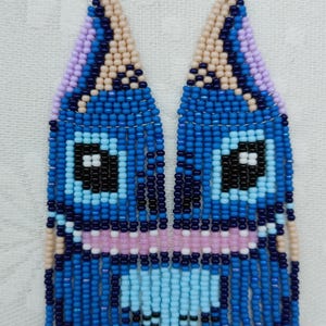 May include: Handmade beaded earrings in a whimsical design. The earrings feature a blue, black, and light blue face-like pattern. The top portion is a triangle shape with a lavender and tan color. The bottom portion has a fringe design.