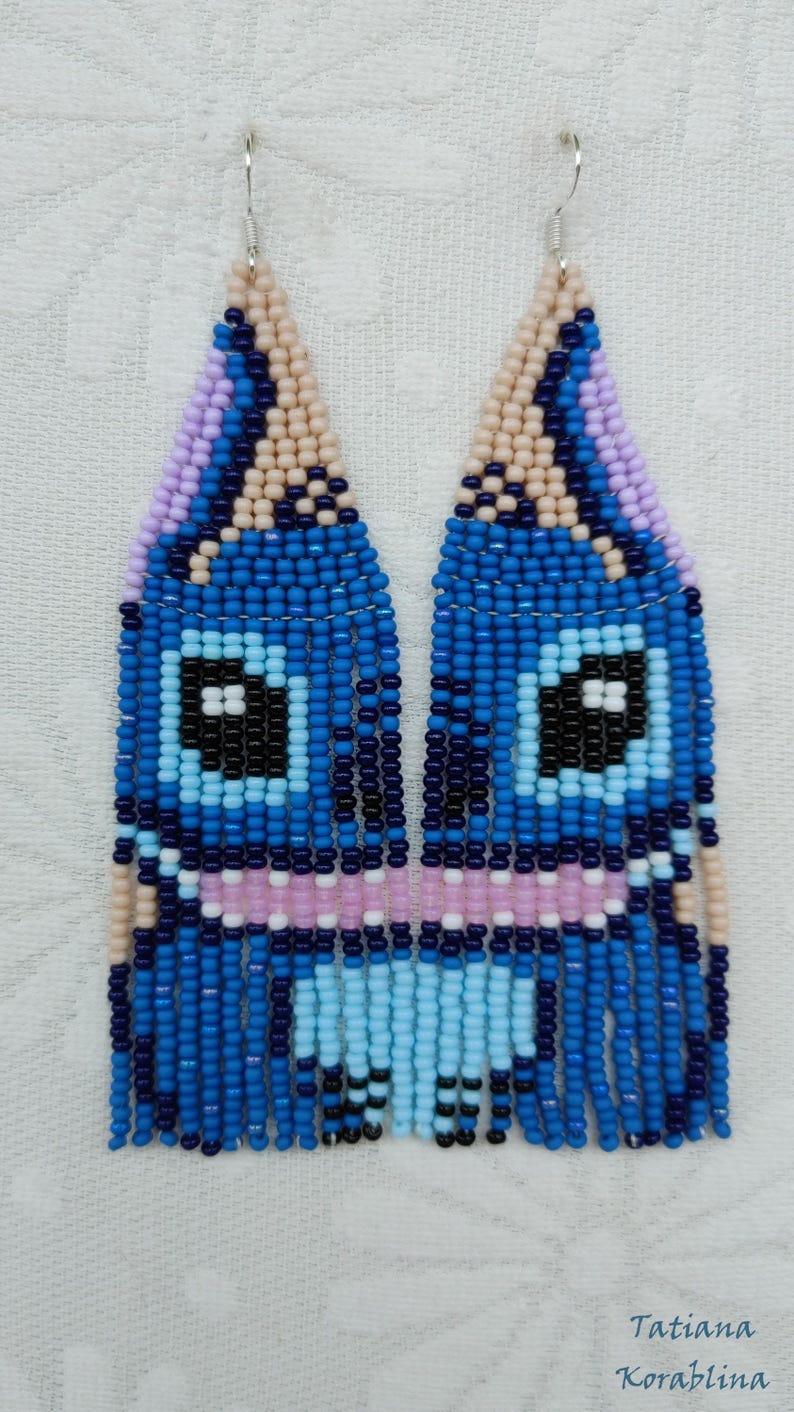 May include: Handmade beaded earrings in a whimsical design. The earrings feature a blue, black, and light blue face-like pattern. The top portion is a triangle shape with a lavender and tan color. The bottom portion has a fringe design.