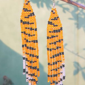 May include: A pair of long, beaded earrings with a tiger stripe pattern in orange and black. The earrings are hanging from a branch.