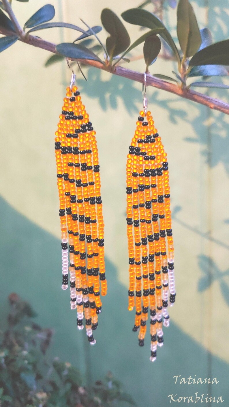 May include: A pair of long, beaded earrings with a tiger stripe pattern in orange and black. The earrings are hanging from a branch.