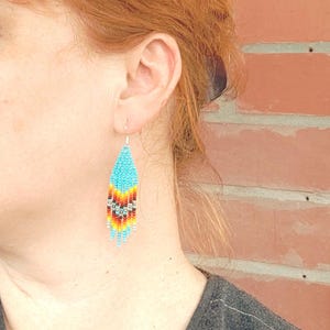 May include: A pair of turquoise, red, orange and yellow beaded earrings. The earrings are teardrop shaped and hang from silver hooks.