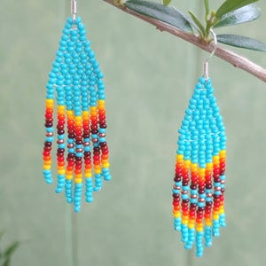 May include: A pair of turquoise beaded earrings with red, orange and yellow accents. The earrings are hanging from a branch.