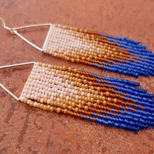 May include: A pair of long, beaded earrings with a gradient of white, beige, brown, and blue beads. The earrings are attached to silver triangle-shaped hoops.