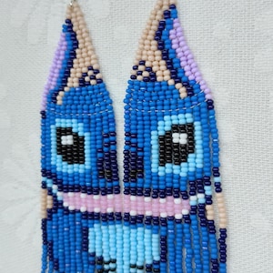 May include: Handmade beaded earrings featuring a blue cartoon character design. The earrings have a light blue, dark blue, pink, black, and beige color scheme. The earrings are made with small beads and have a fringe at the bottom.