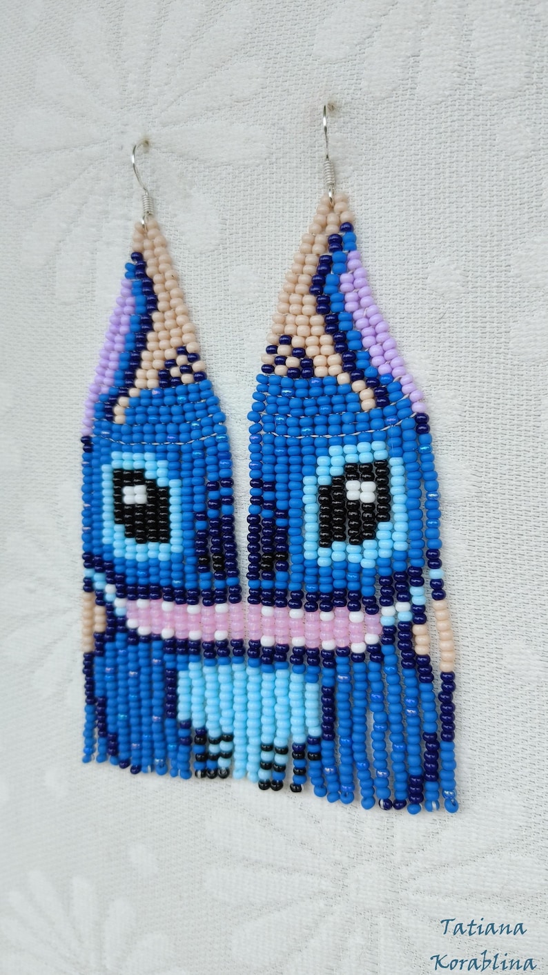 May include: Handmade beaded earrings featuring a blue cartoon character design. The earrings have a light blue, dark blue, pink, black, and beige color scheme. The earrings are made with small beads and have a fringe at the bottom.