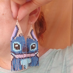 May include: Handmade beaded earrings featuring a blue cartoon character design. The earrings have a rectangular shape with a fringe at the bottom. The design incorporates shades of blue, pink, and beige.