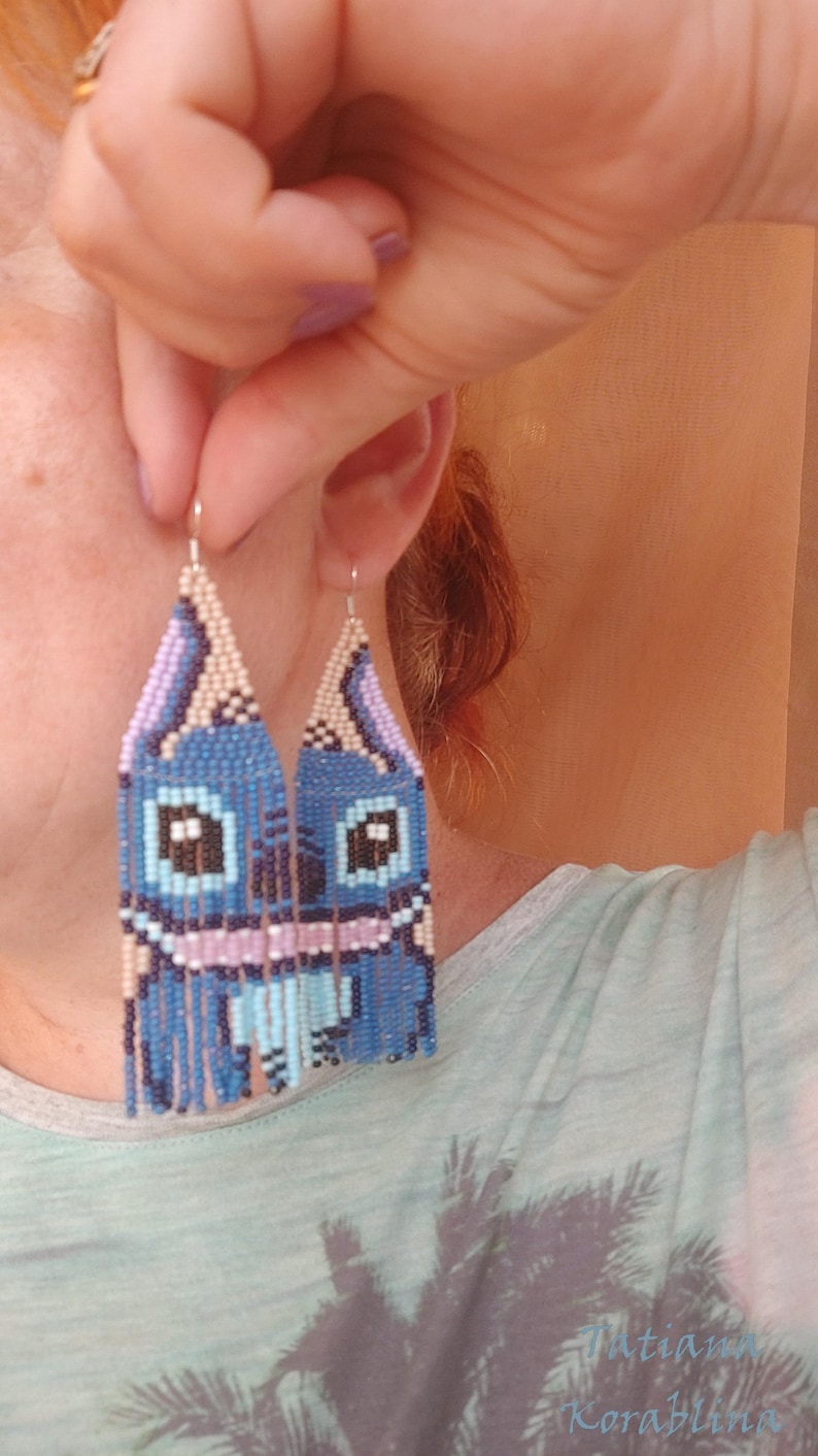 May include: Handmade beaded earrings featuring a blue cartoon character design. The earrings have a rectangular shape with a fringe at the bottom. The design incorporates shades of blue, pink, and beige.