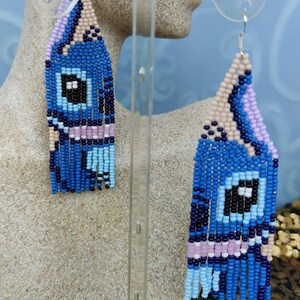 May include: Handmade beaded earrings in a geometric design. The earrings feature a blue, black, and white color scheme with fringe detailing. The earrings are displayed on a mannequin head. The earrings are a unique accessory.