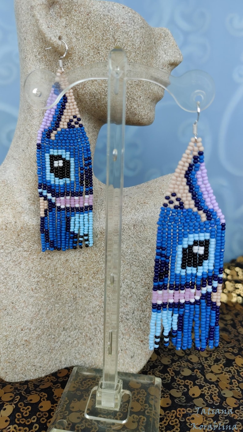 May include: Handmade beaded earrings in a geometric design. The earrings feature a blue, black, and white color scheme with fringe detailing. The earrings are displayed on a mannequin head. The earrings are a unique accessory.