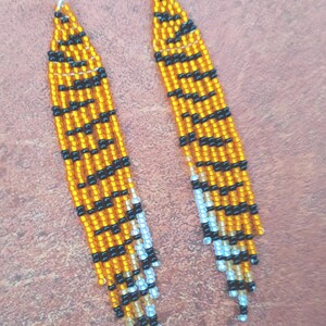 May include: A pair of long, beaded earrings with a tiger print design. The earrings are made with orange, black, and white beads. The earrings are hanging from silver hooks.