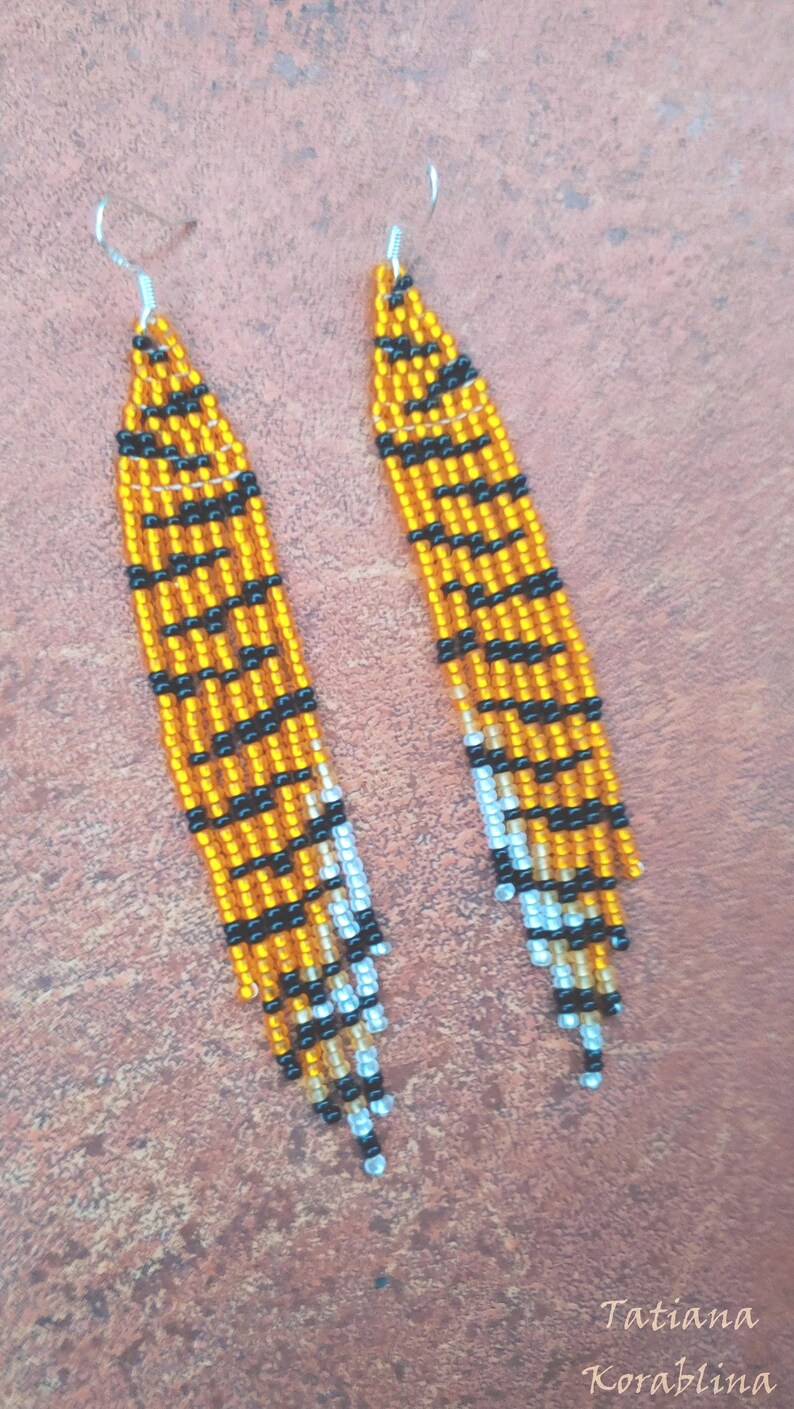 May include: A pair of long, beaded earrings with a tiger print design. The earrings are made with orange, black, and white beads. The earrings are hanging from silver hooks.