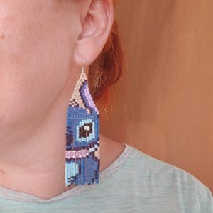 May include: Handmade beaded earrings in a blue, purple, and beige design. The earrings feature a rectangular shape with a fringe at the bottom. The design appears to be a cartoon character. The earrings are hanging from a silver hook.