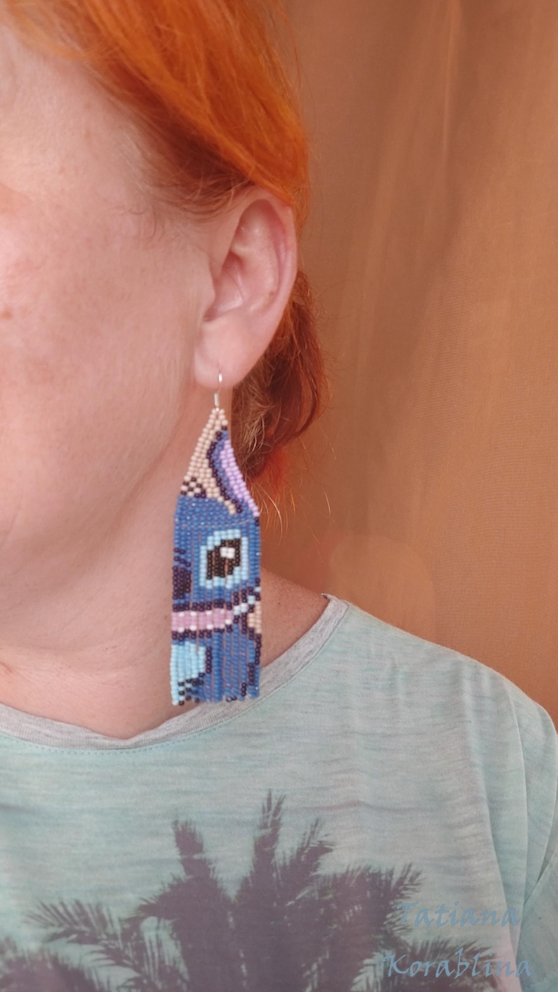 May include: Handmade beaded earrings in a blue, purple, and beige design. The earrings feature a rectangular shape with a fringe at the bottom. The design appears to be a cartoon character. The earrings are hanging from a silver hook.