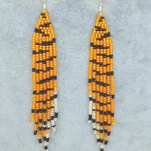 May include: A pair of long, orange and black beaded earrings with a white fringe. The earrings are made with small beads and have a tiger stripe pattern.