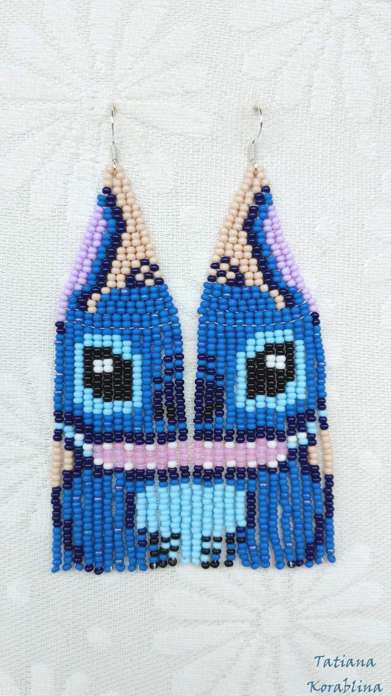 May include: Handmade beaded earrings in a whimsical design. The earrings feature a blue, light blue, pink, and beige color scheme. The design includes a face-like pattern with black square eyes and a fringe at the bottom. The earrings are suspended from silver hooks.