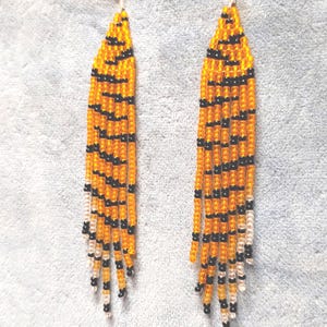May include: A pair of long, beaded earrings with a tiger stripe pattern in orange and black. The earrings are made with small, round beads and have a silver hook closure.