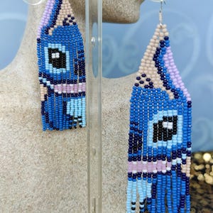 May include: Blue beaded earrings with a geometric design. The earrings feature a combination of blue, black, white, and pink beads, with a fringe at the bottom. The earrings are displayed on a clear stand.