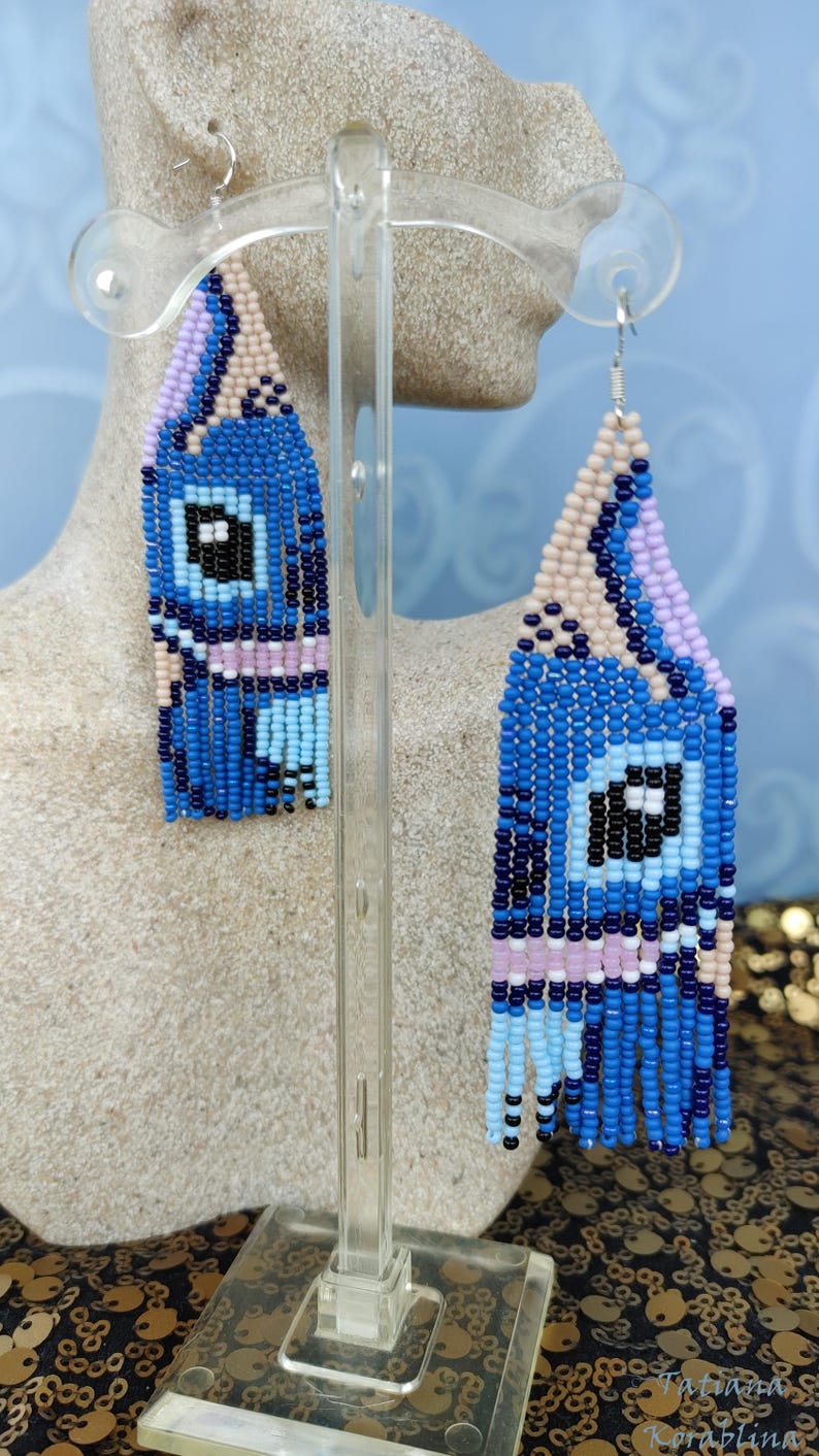 May include: Blue beaded earrings with a geometric design. The earrings feature a combination of blue, black, white, and pink beads, with a fringe at the bottom. The earrings are displayed on a clear stand.