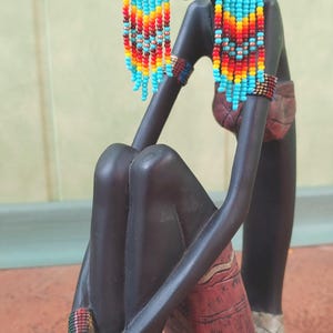 May include: A black figurine of a woman wearing long, beaded earrings with turquoise, orange, yellow, and red beads. The earrings are dangling and have a fringe-like appearance. The figurine is sitting with its legs crossed and has a textured, red and brown skirt.
