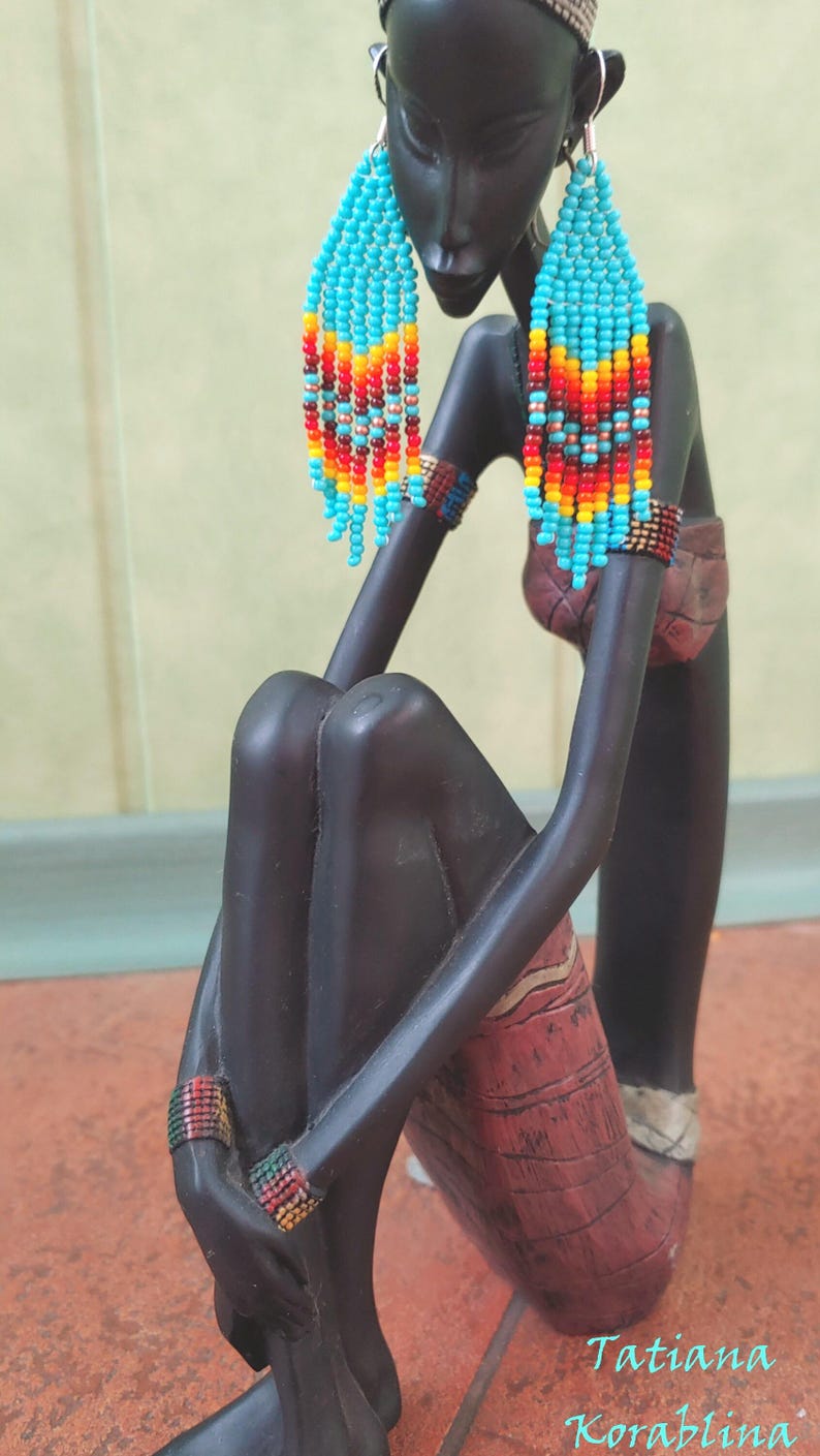 May include: A black figurine of a woman wearing long, beaded earrings with turquoise, orange, yellow, and red beads. The earrings are dangling and have a fringe-like appearance. The figurine is sitting with its legs crossed and has a textured, red and brown skirt.